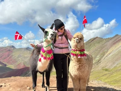 TourCompass-kollegan Iida står med två flaggor och en alpaka på toppen av Rainbow Mountain i Peru.