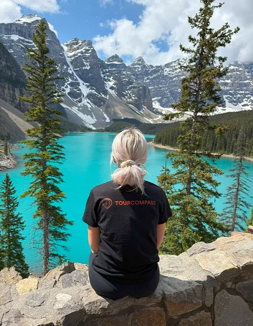 Kvinna med TourCompass T-shirt blickar ut över en turkosblå sjö och snötäckta berg i Kanada